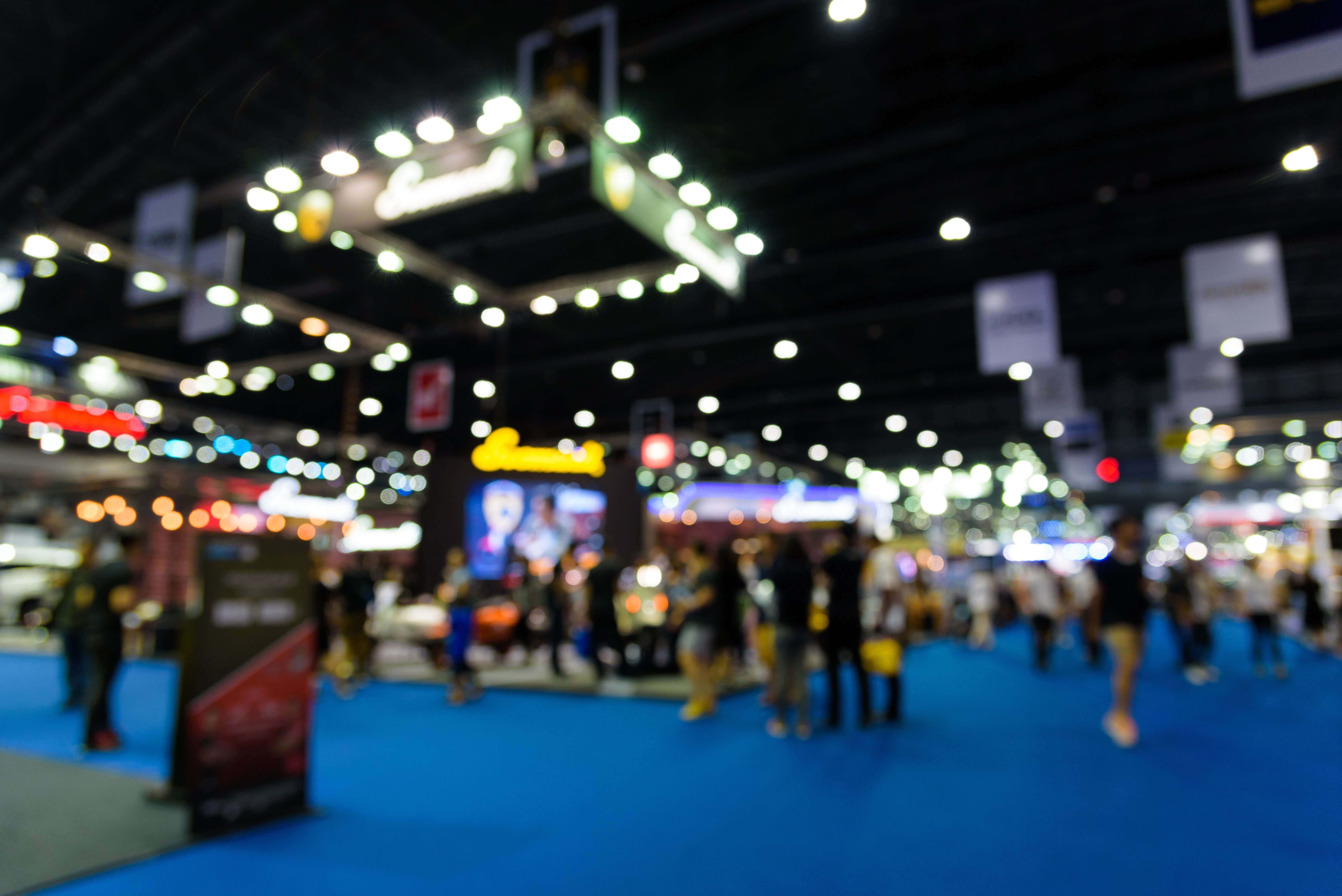 A blurred image of a busy convention or trade show with people walking and visiting booths under bright lights in a large indoor venue with blue carpeting.