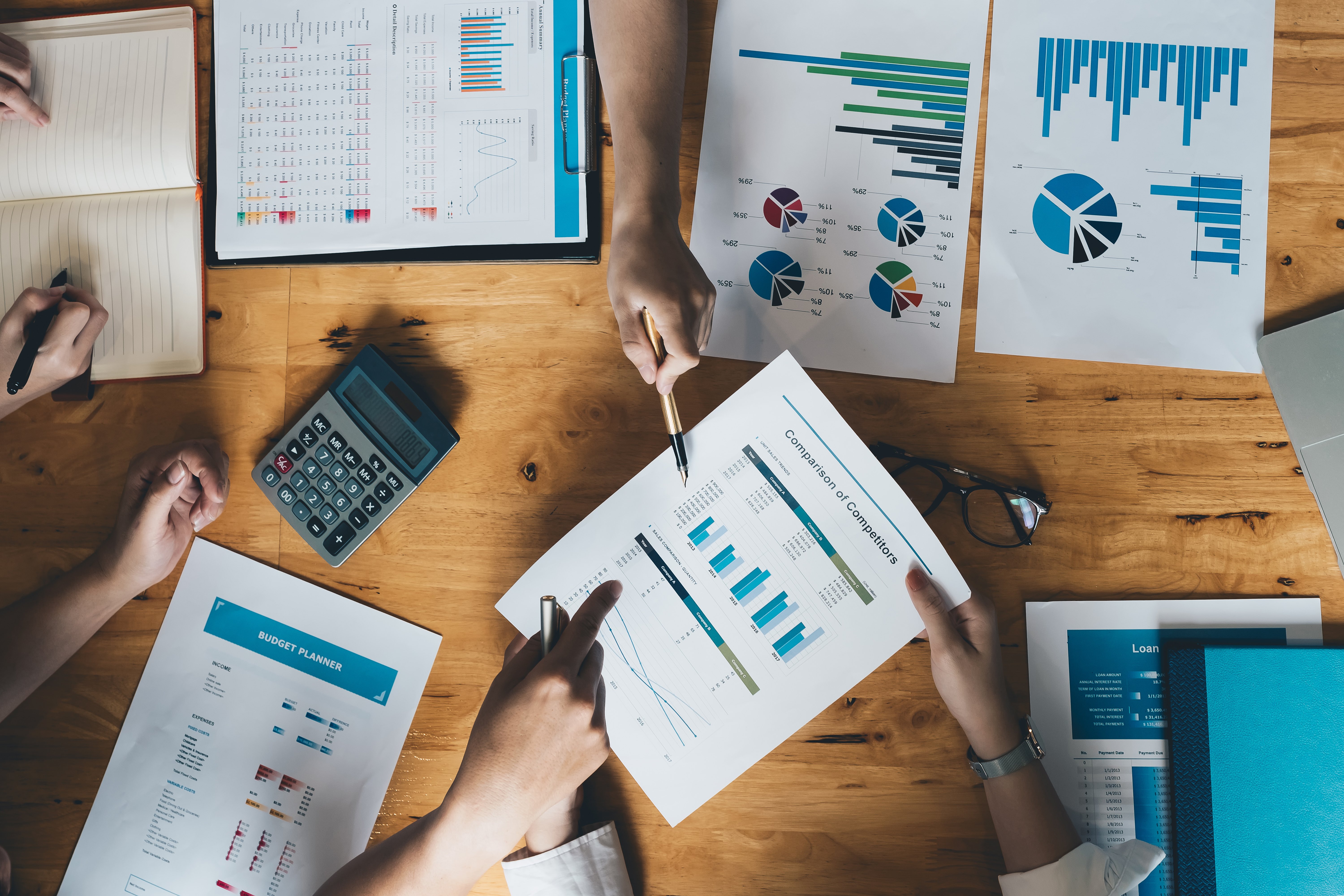 A group of people collaborating on financial and marketing analysis at a wooden table covered with charts, graphs, and reports showing data comparisons, with calculators, notebooks, and pens in use during the discussion.