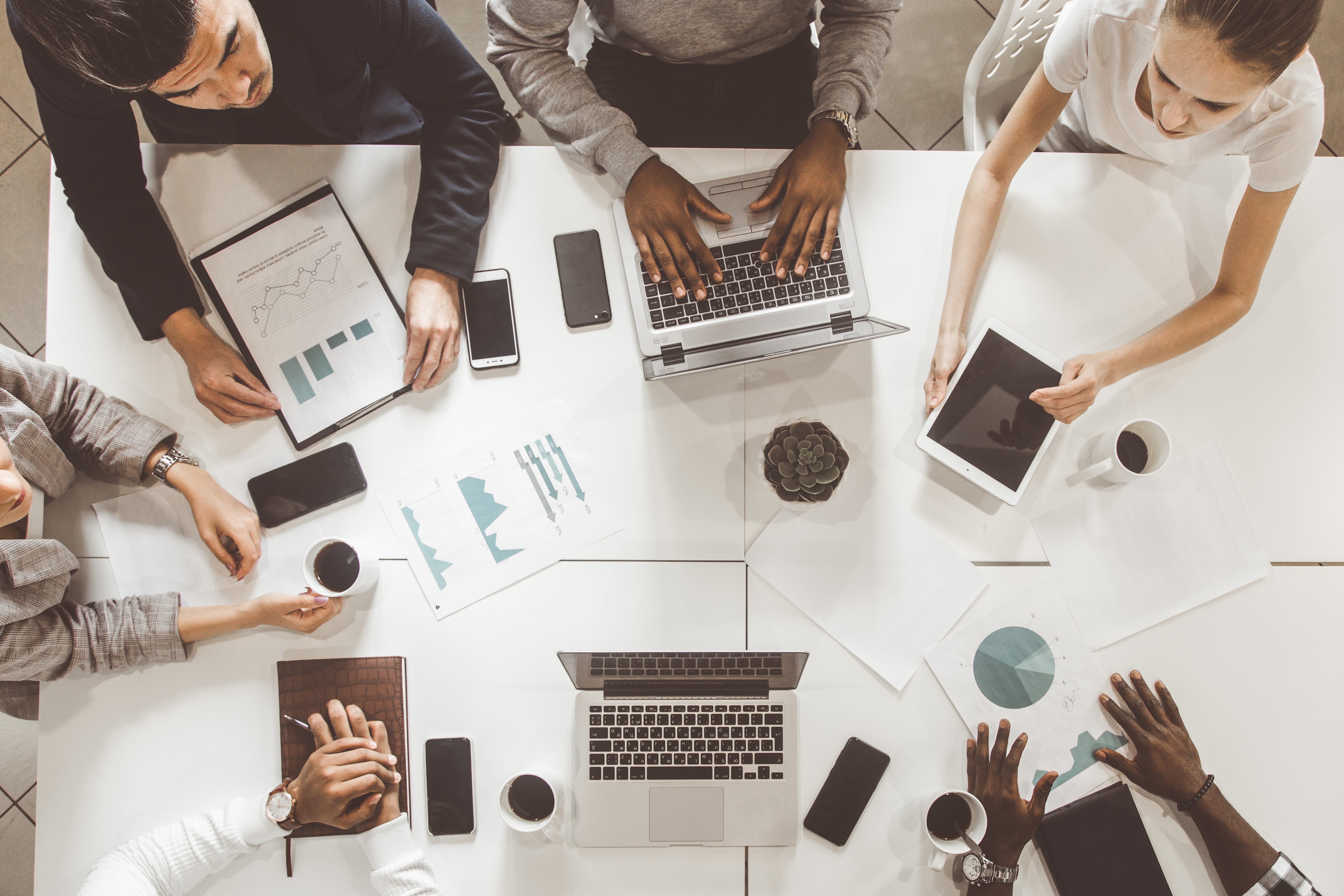A diverse team of professionals collaborating around a white table with laptops, tablets, smartphones, charts, and coffee cups, viewed from above. The group appears to be discussing data and project plans in a modern office setting.