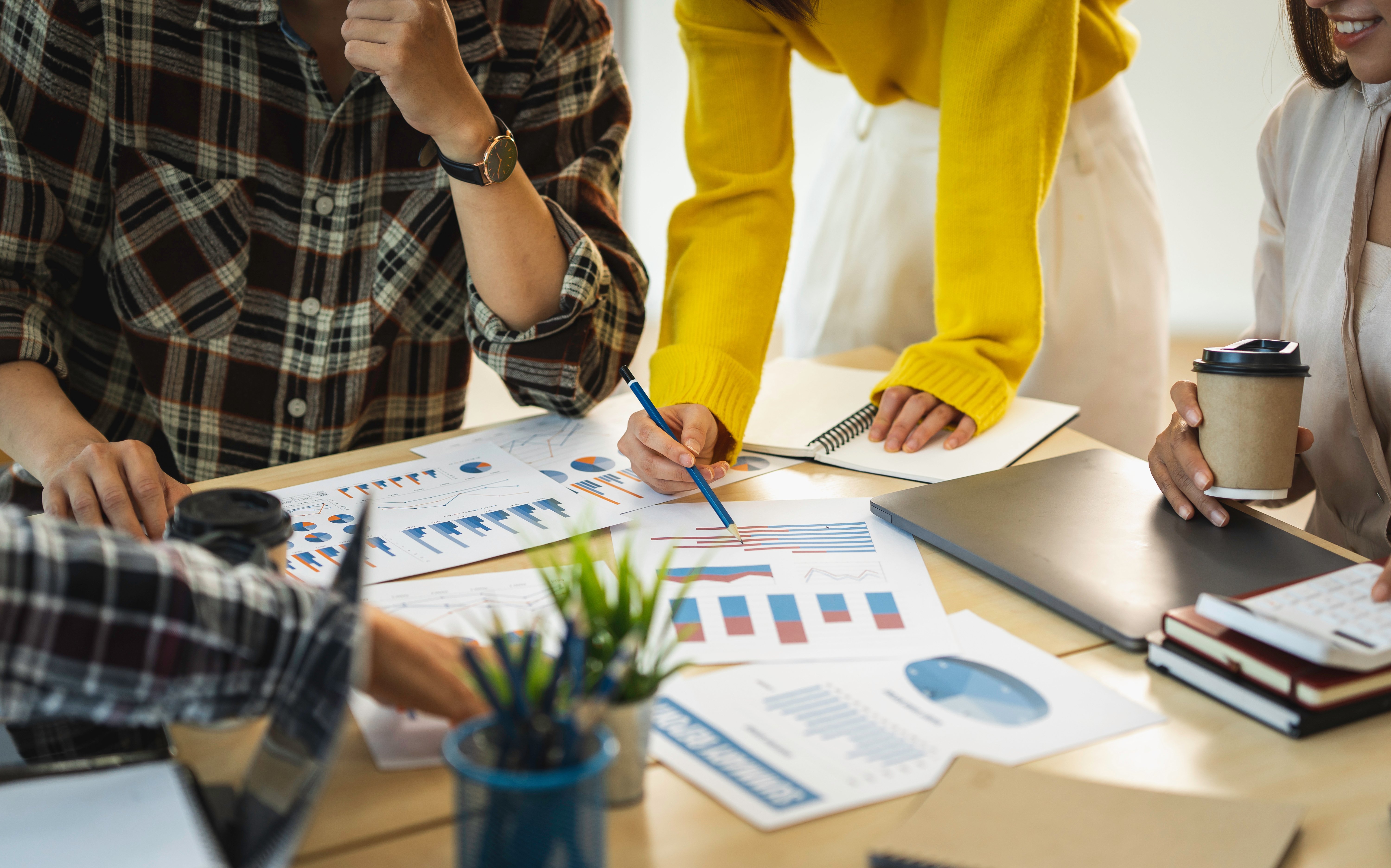 Close-up of a diverse team collaborating at a wooden table, reviewing printed charts and graphs with colorful bar and pie diagrams. One person in a plaid shirt is thinking while another in a bright yellow sweater points to a document with a pencil. Other team members hold a coffee cup and a calculator, surrounded by a laptop, notebooks, and a small potted plant.