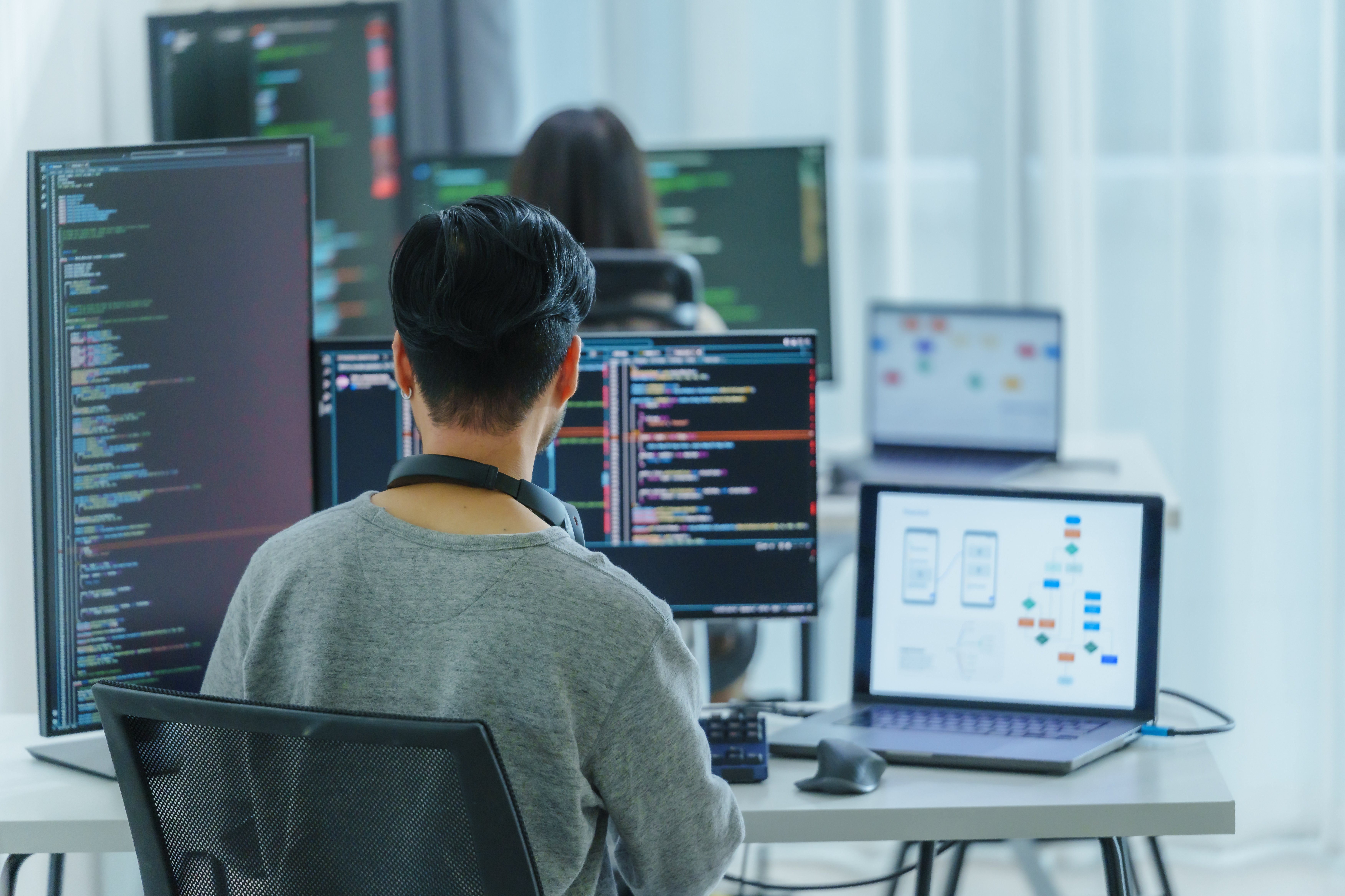 A software developer sitting at a desk surrounded by multiple monitors displaying code and a laptop showing a flowchart or app design, working in a modern, bright office environment with other programmers in the background.