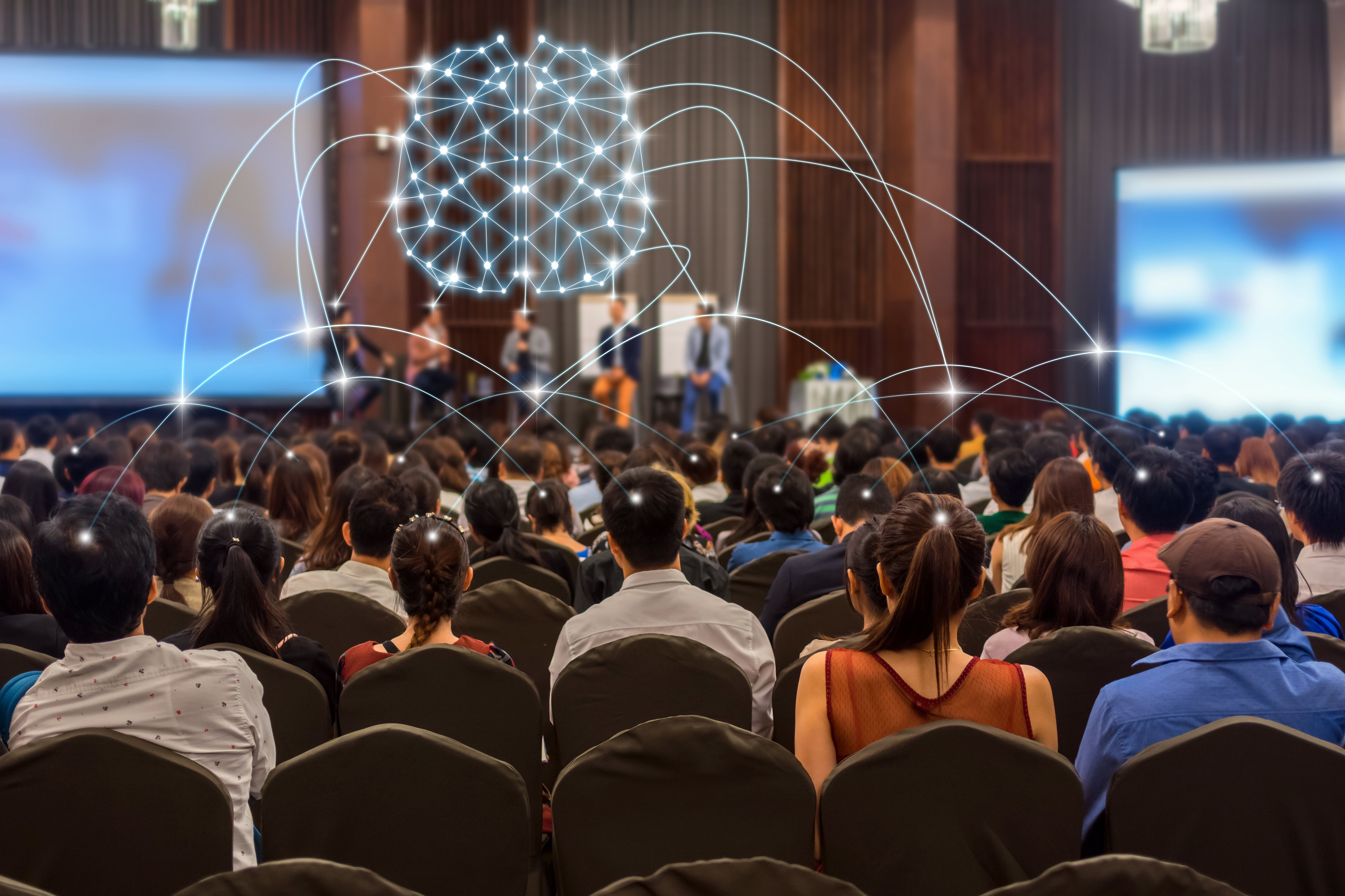 A large audience attending a conference or seminar, listening to speakers on stage, with a glowing digital illustration of a connected brain symbolizing artificial intelligence and data networking overlayed above the crowd.