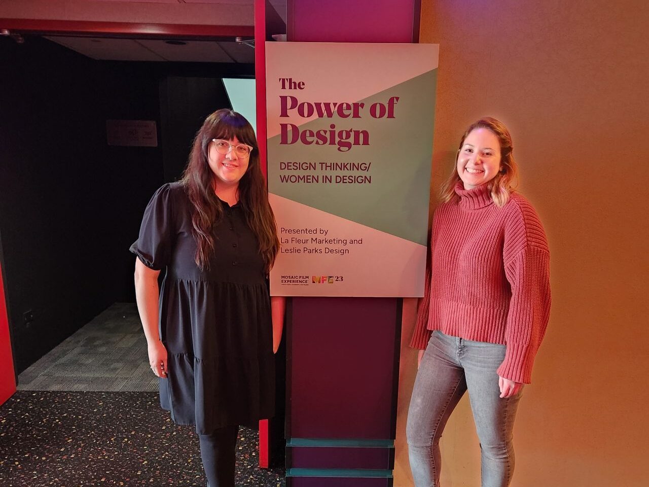 Two women posing next to a sign about the power of design at an event.