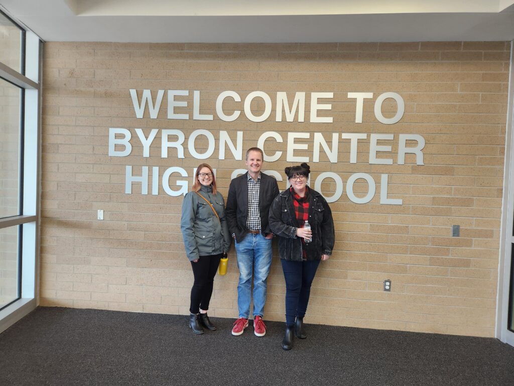 Two women and one man standing in front of wood slat wall and a 'Welcome to Byron Center High School' sign.