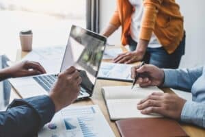 Three professionals seated at a table, discussing how to grow a law firm. One person types on a laptop displaying graphs, while another writes notes, and a third reviews printed charts. Coffee cups and a bright office setting enhance the collaborative atmosphere.