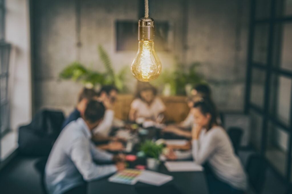 Scalable Marketing Systems for Professional Services Firms 3 Glowing Edison light bulb hanging in front of a blurred team meeting around a conference table